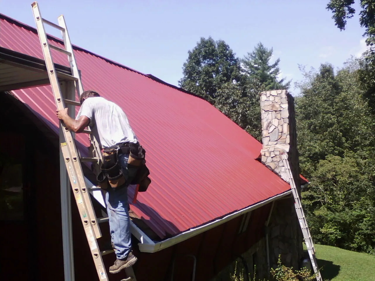 Roofer on a ladder beside a red metal roof and stone chimney — Soffit & Fascia Repair by Roofing Brothers in Hagerman